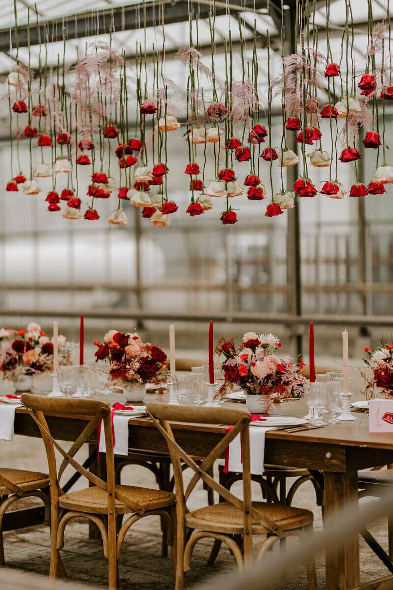 Table et chaises en bois décorées pour un mariage. Fleurs rouges et roses suspendus à une immense verrière flottants dans les airs. Scène imaginée par Laura, pour le workshop photographie de mariage.