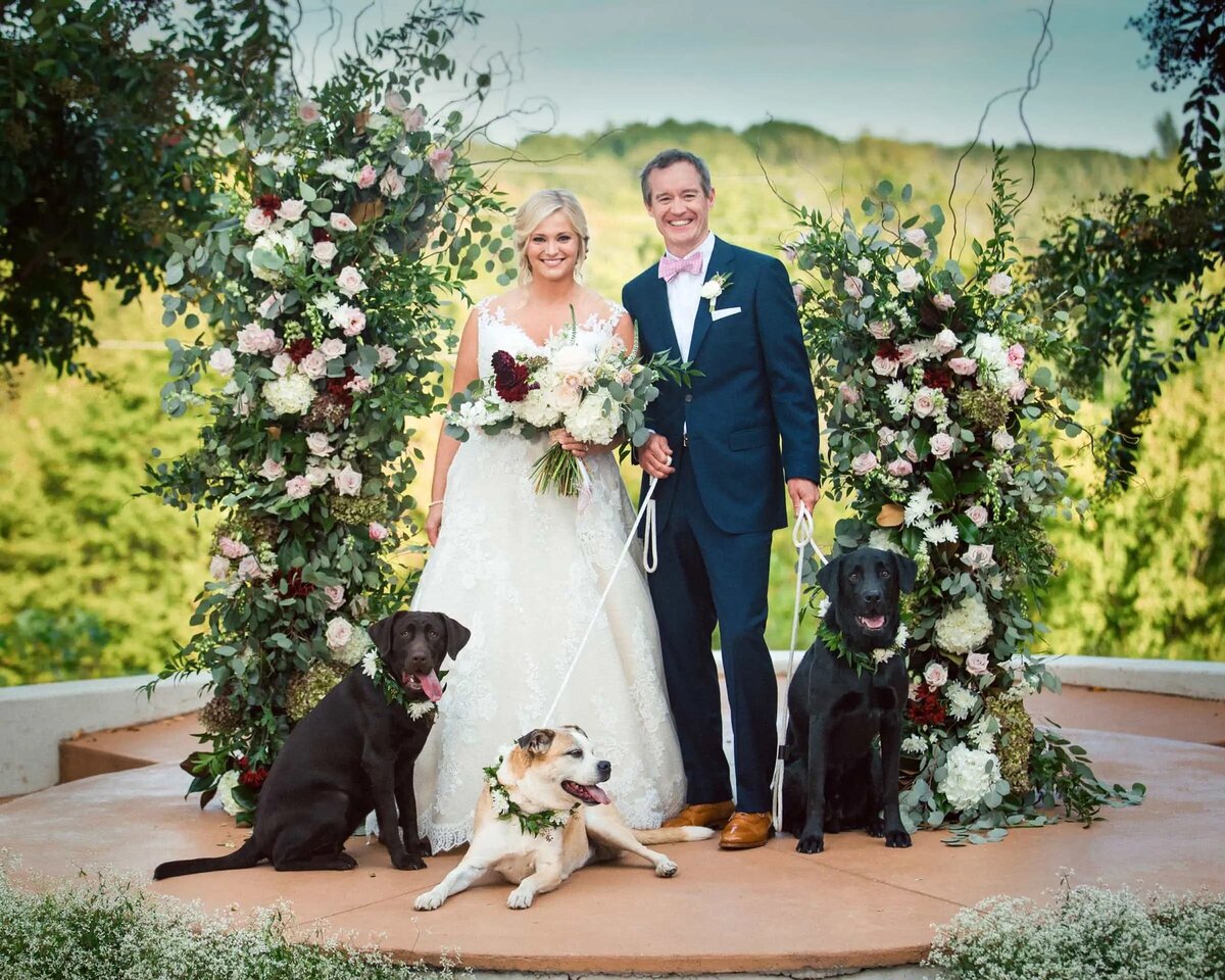 A beaming bride and groom stand with their dogs amidst a floral archway,