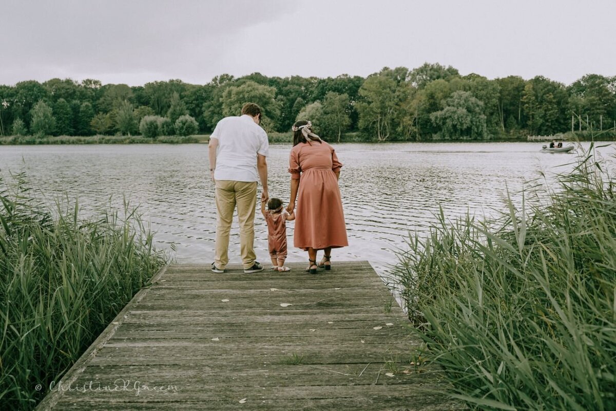 Parents holding toddler at lakeside – Mother and father gently hold their little girl’s hands as they walk together to the end of a wooden pier surrounded by lush greenery.