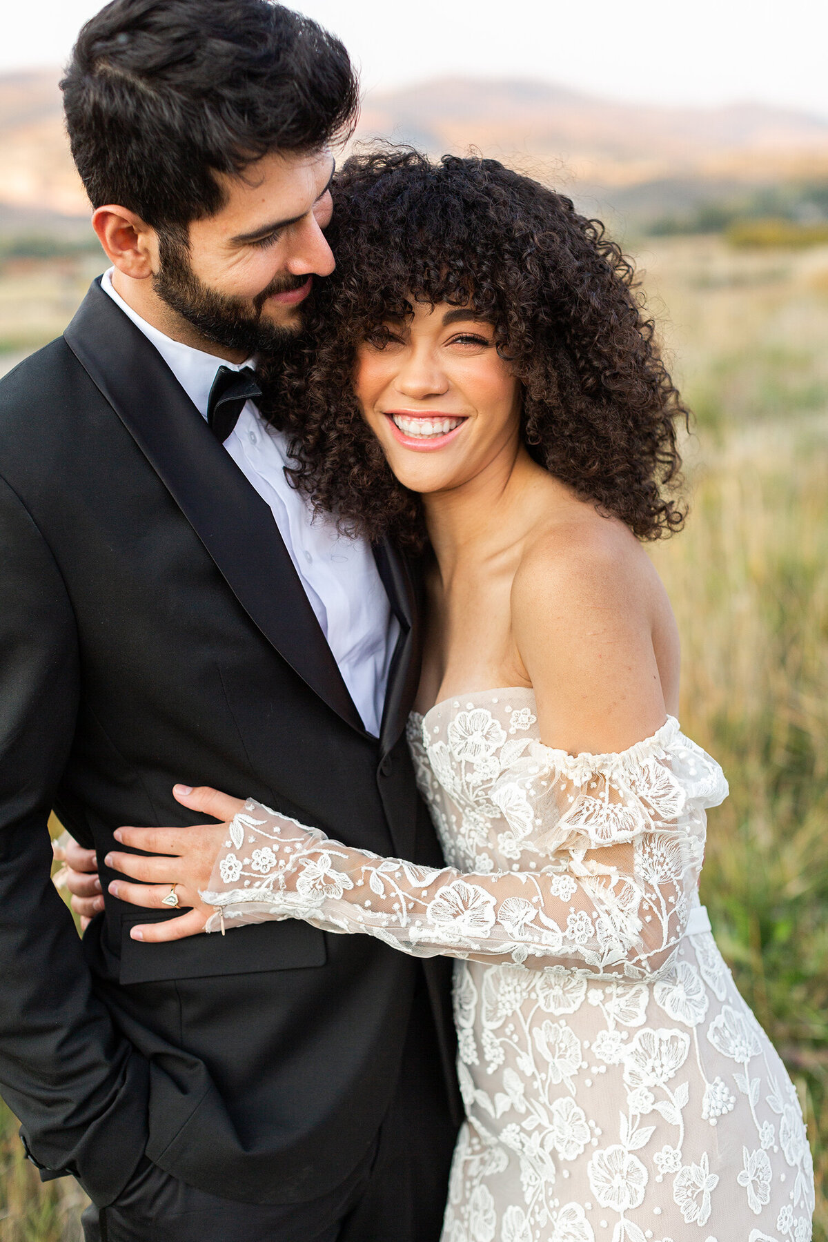 man wearing a black tuxedo, affectionately holding the woman whose smiling joyfully in an off-shoulder white lace wedding dress