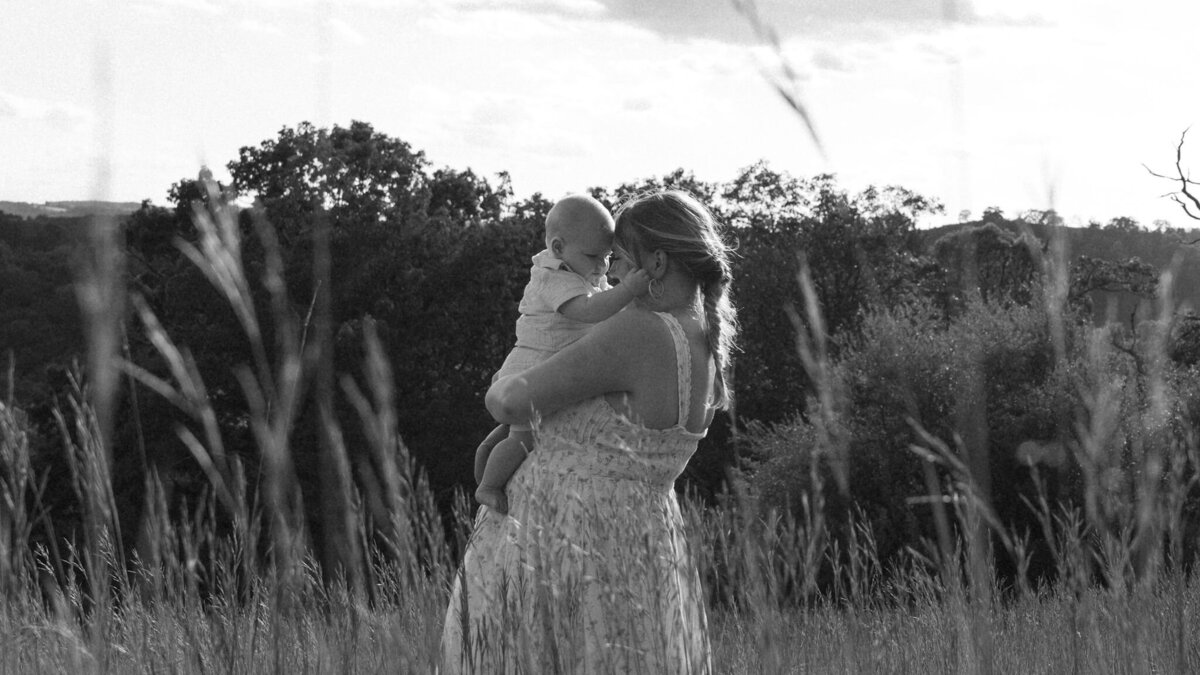 black and white photo of a mom and with her baby holding her face gently while standing in a field of tall grass.