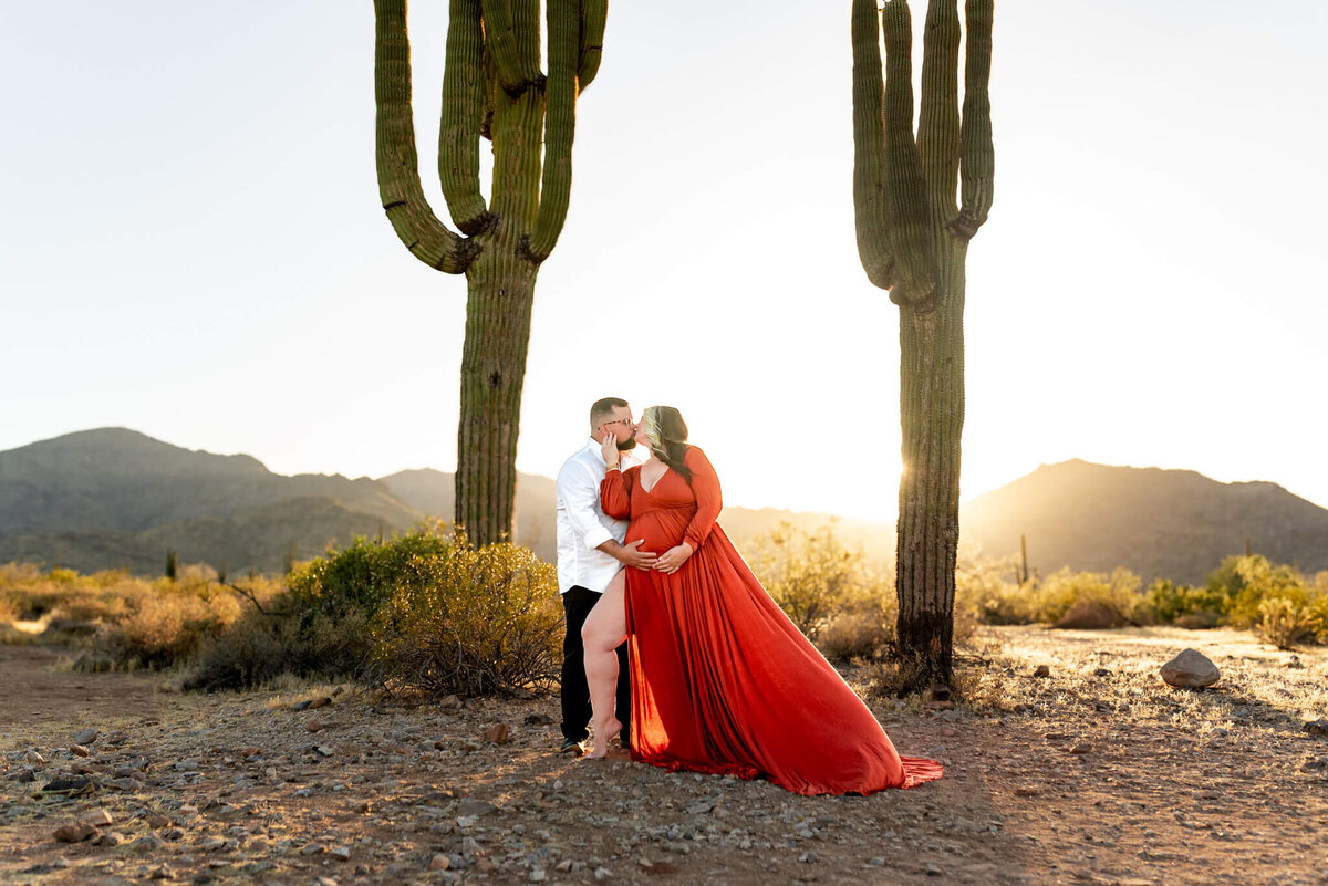 Expecting parents share a kiss at the last bit of sunset, between 2 saguaros