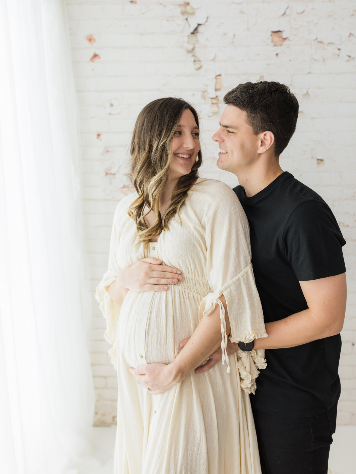 husband and pregnant  wife in cream maternity dress holding baby bump for maternity photoshoot