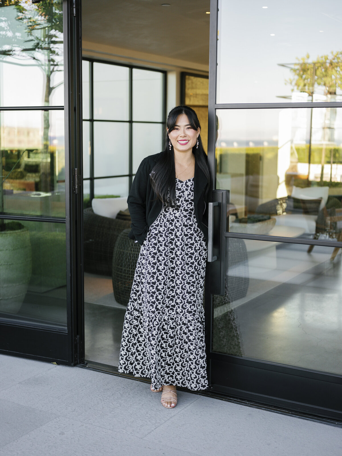 Asian woman smiling in patterned dress and black jacket at office doorway during NYC brand shoot.