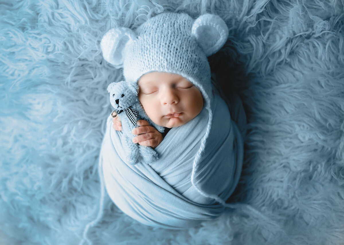 A newborn baby sleeps in a blue swaddle and bear bonnet while cuddling matching stuffed bear