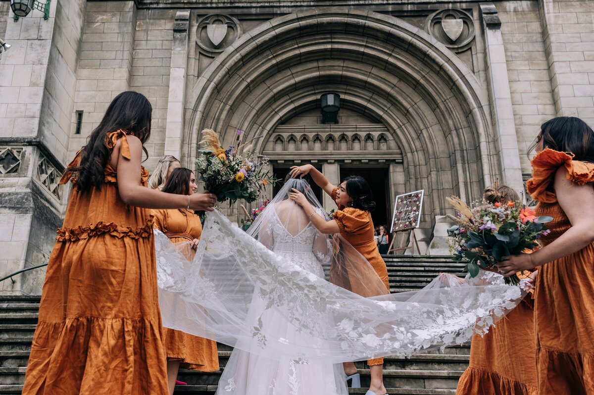 bridesmaids in burnt orange adjust brides veil on steps of st pauls cathedral in dunedin