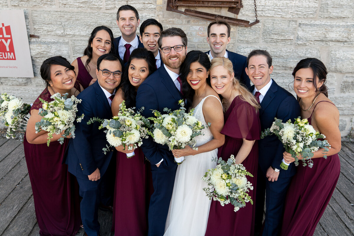 Bride and groom snuggle with wedding party in front of Mill City Museum in Minneapolis, Minnesota.