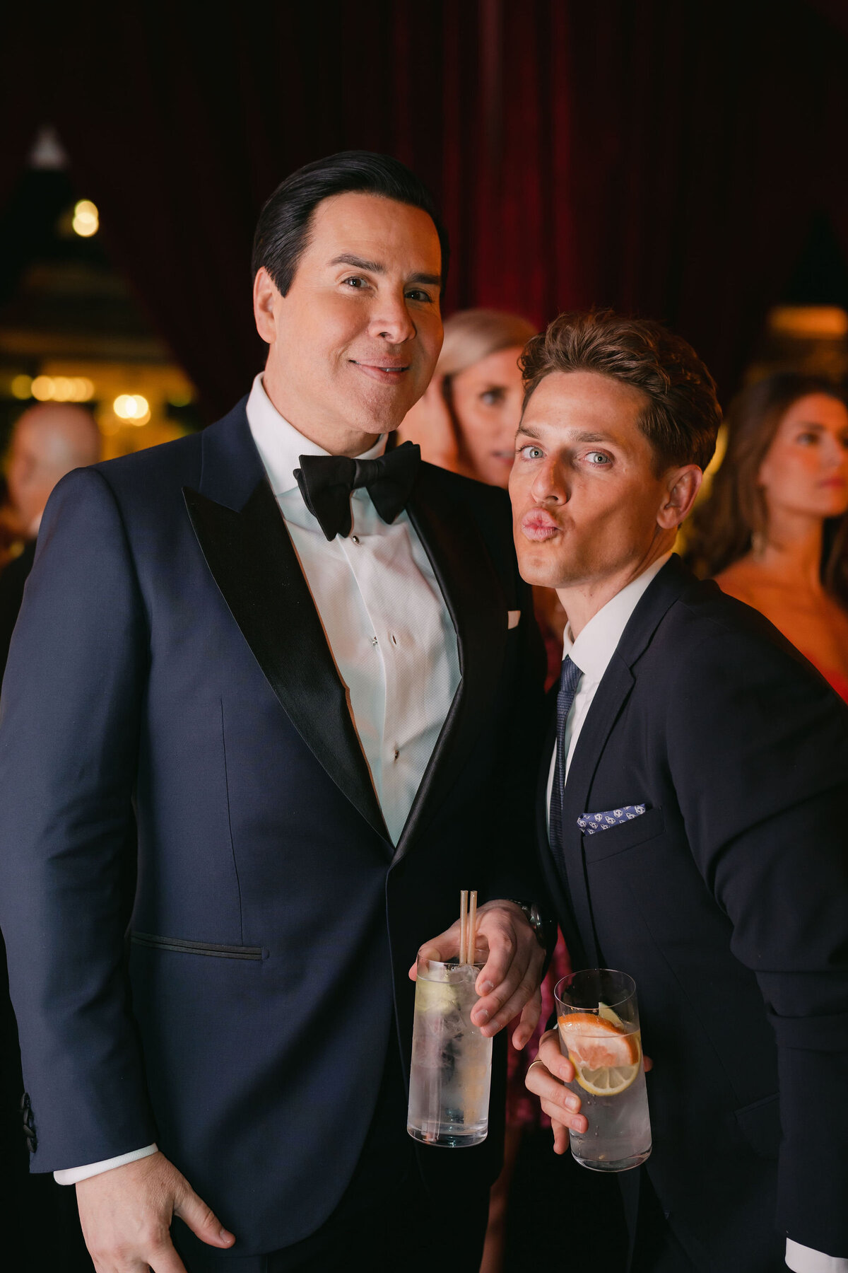 Two men dressed in formal suits pose at an event. The man on the left, in a tuxedo, smiles while holding a drink with lemon garnish. The man on the right, also in a suit, playfully puckers his lips while holding a drink with a grapefruit slice. Other attendees are in the background.