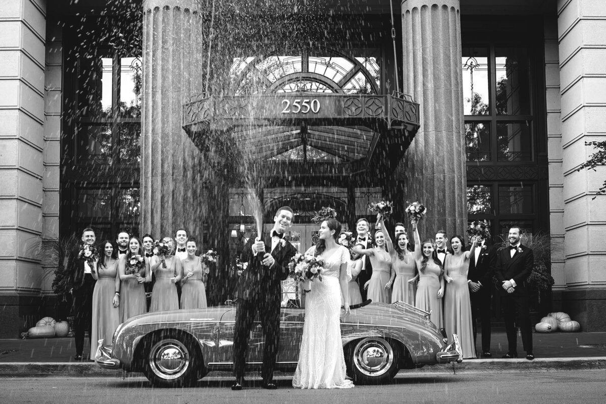 Black and white portrait of bride and groom popping champagne by a vintage car in front of the wedding party