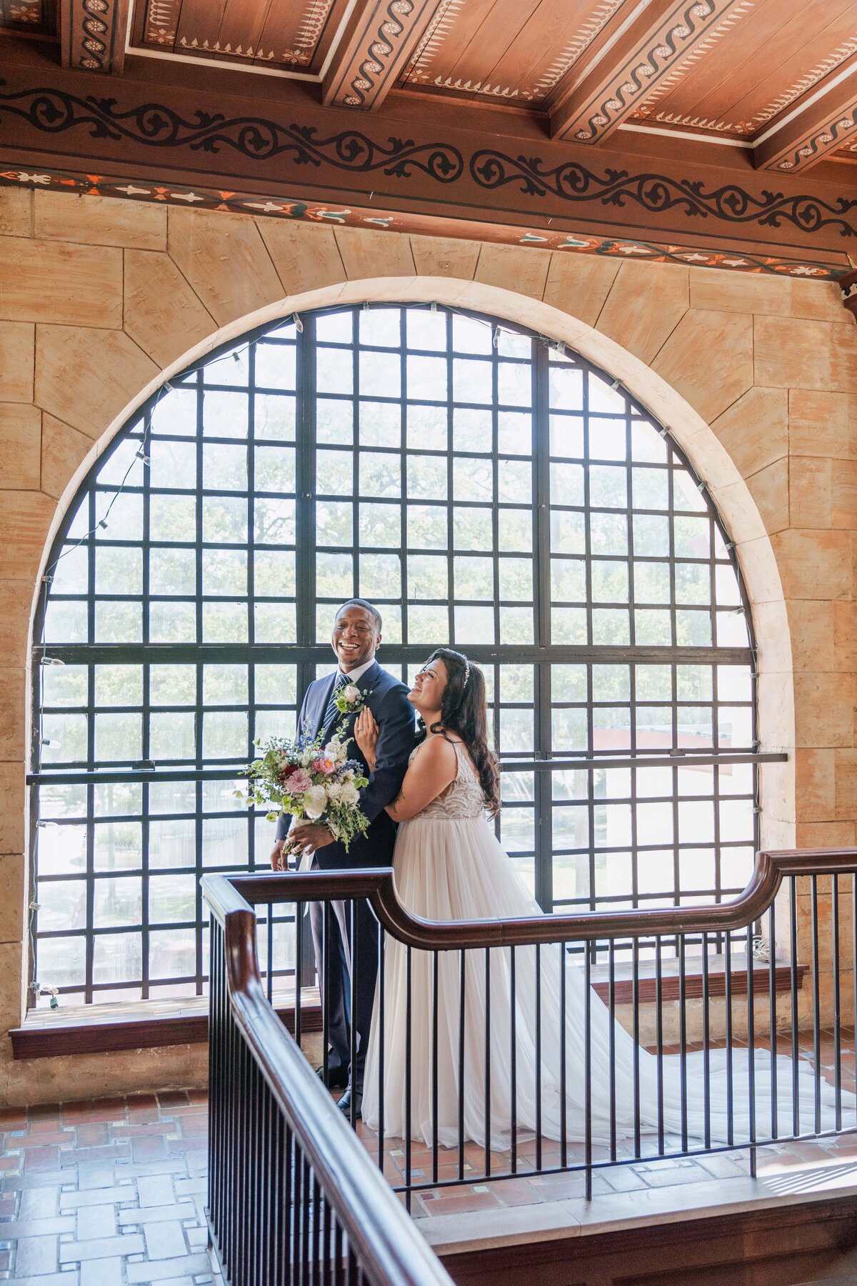 A joyful first look between a bride and groom at The Treasury on the Plaza in St. Augustine, Florida. The couple stands in front of the venue’s iconic arched window, bathed in natural light. The bride holds a romantic bouquet of soft florals while smiling up at her groom, who looks thrilled and emotional. Captured by Phavy Photography in a true-to-color, documentary style that beautifully preserves the genuine emotion and architectural charm of this St. Augustine wedding venue.
