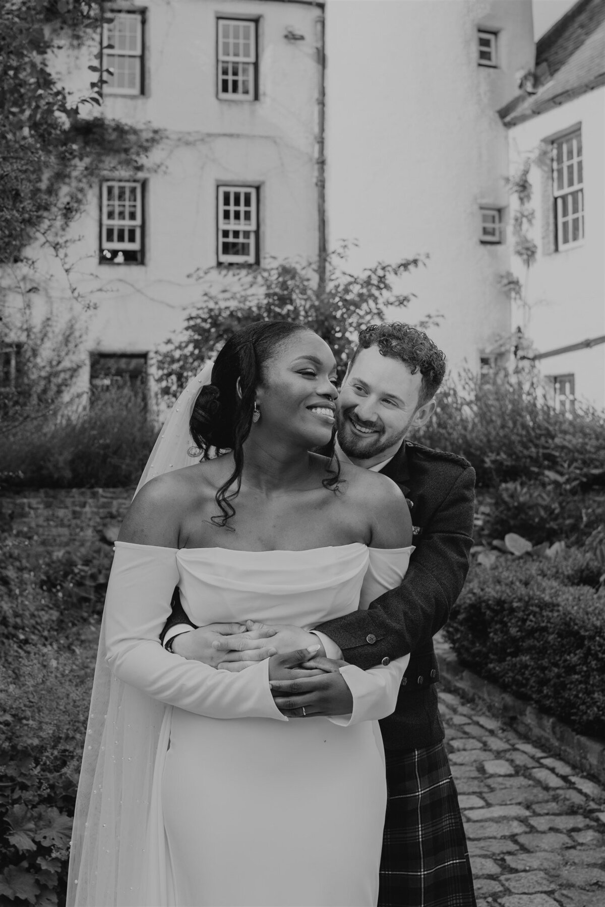 A groom wraps his arms around his wife from behind. They stand in a shaded courtyard and the white exterior of Logie Country House can be seen all around them. They stand on a cobbled surface and pose for wedding photos. The photo is black and white,