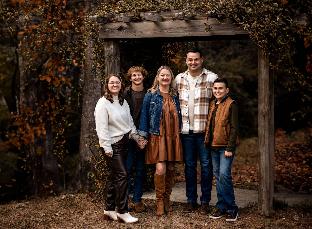 A family wearing fall clothes standing in front of a wooden archway draped with foliage in Fairhope, Alabama