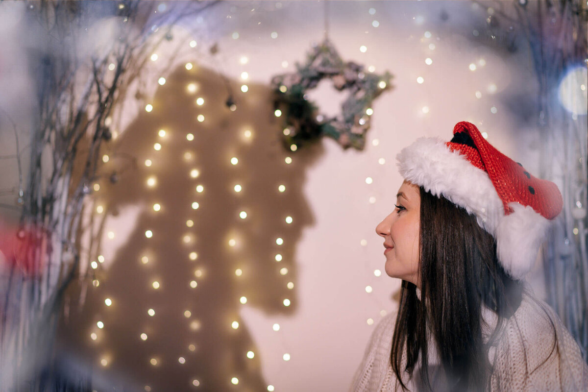 Woman wearing Santa hat admiring Christmas tree lights in Calini Weddings Studio - festive glow and warm holiday atmosphere.