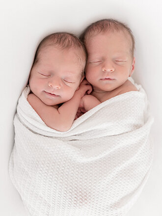 Newborn twins cuddled together in a white wrap on a whiit blanket.
