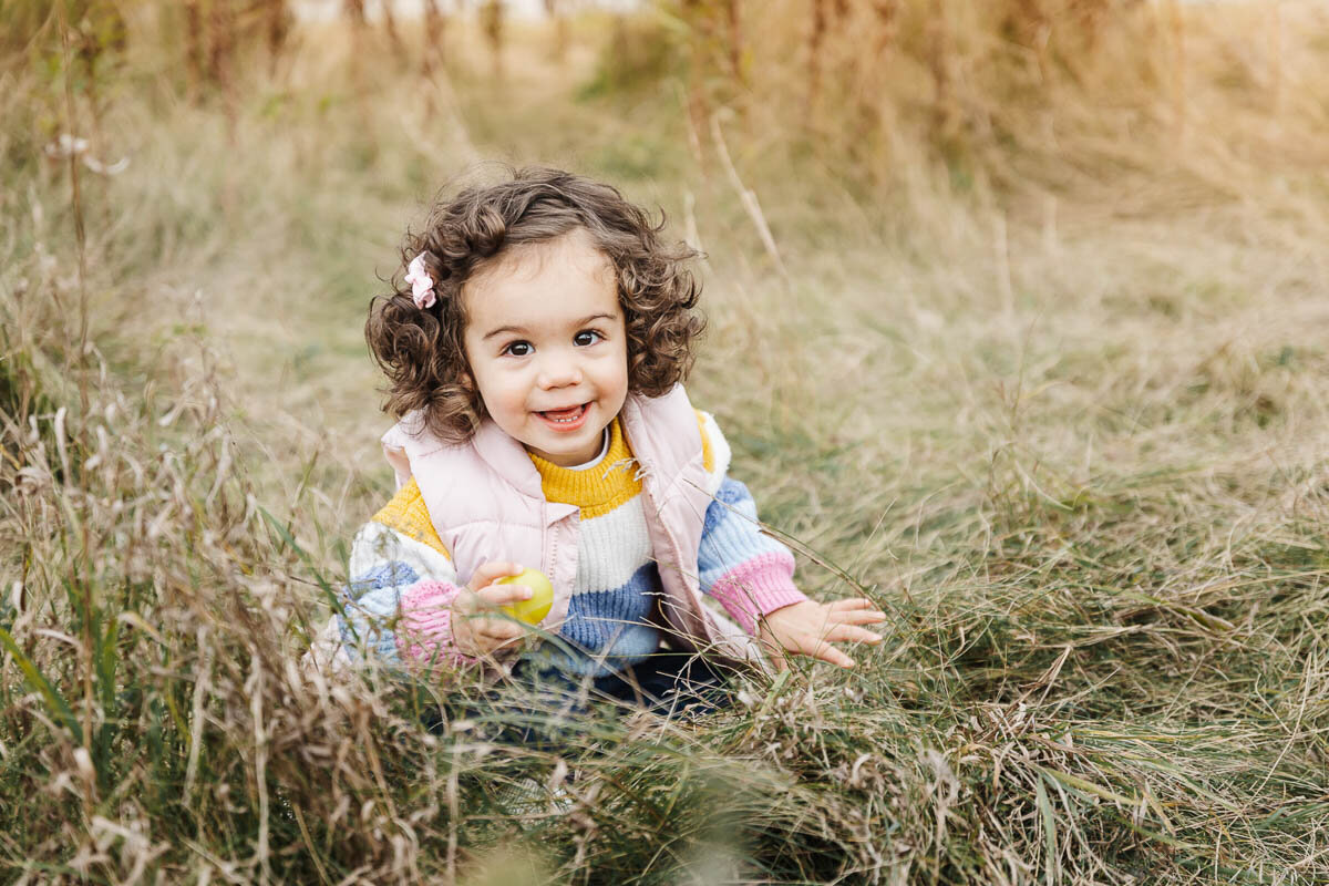 A young child with curly hair, dressed in a colorful sweater and pink vest, sits amidst the tall grass of a serene Toronto park, clutching a yellow ball. It's a perfect moment captured in family photography.