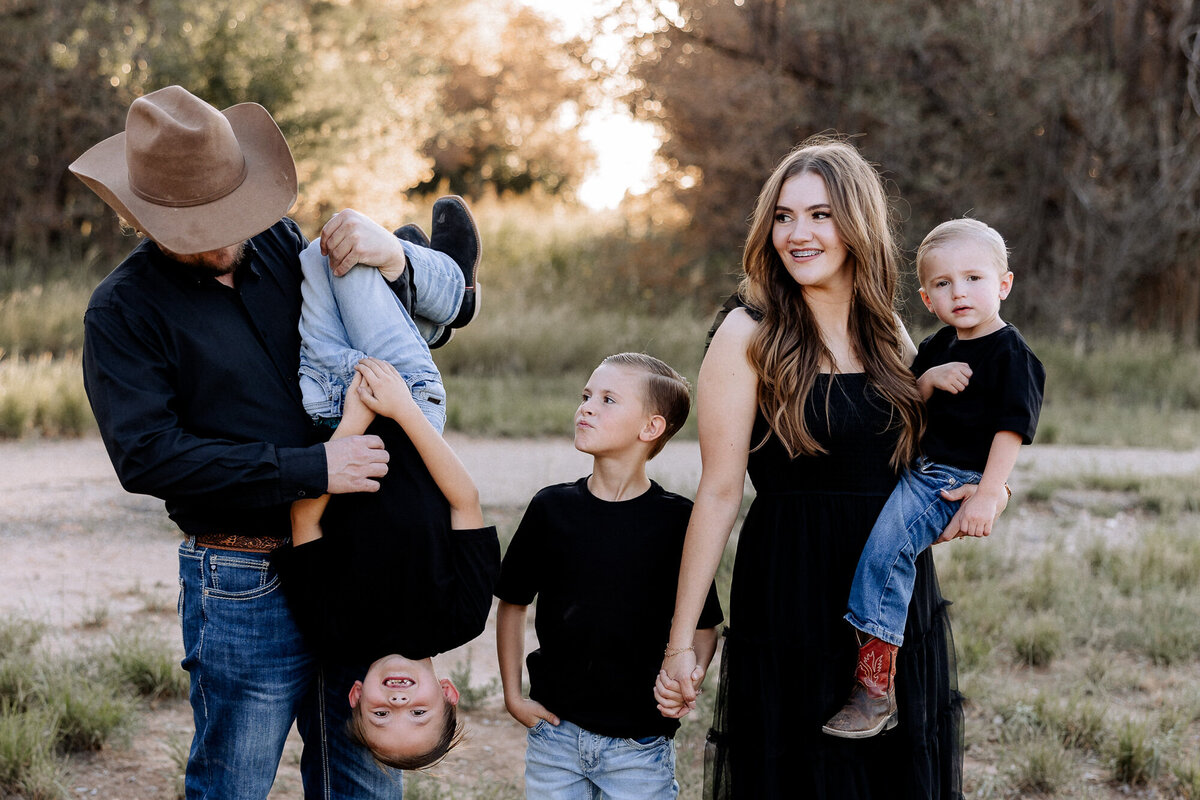 joyful family of five in a grass field with the sun setting behind them. Dad is playing with son holding him upside down. mom smiling over at dad and son playing. Captured by Mary Wiebe Photograpphy