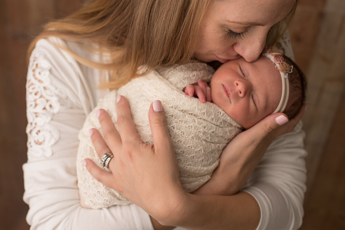 Woman in white holding newborn girl wrapped in white, kissing her cheek