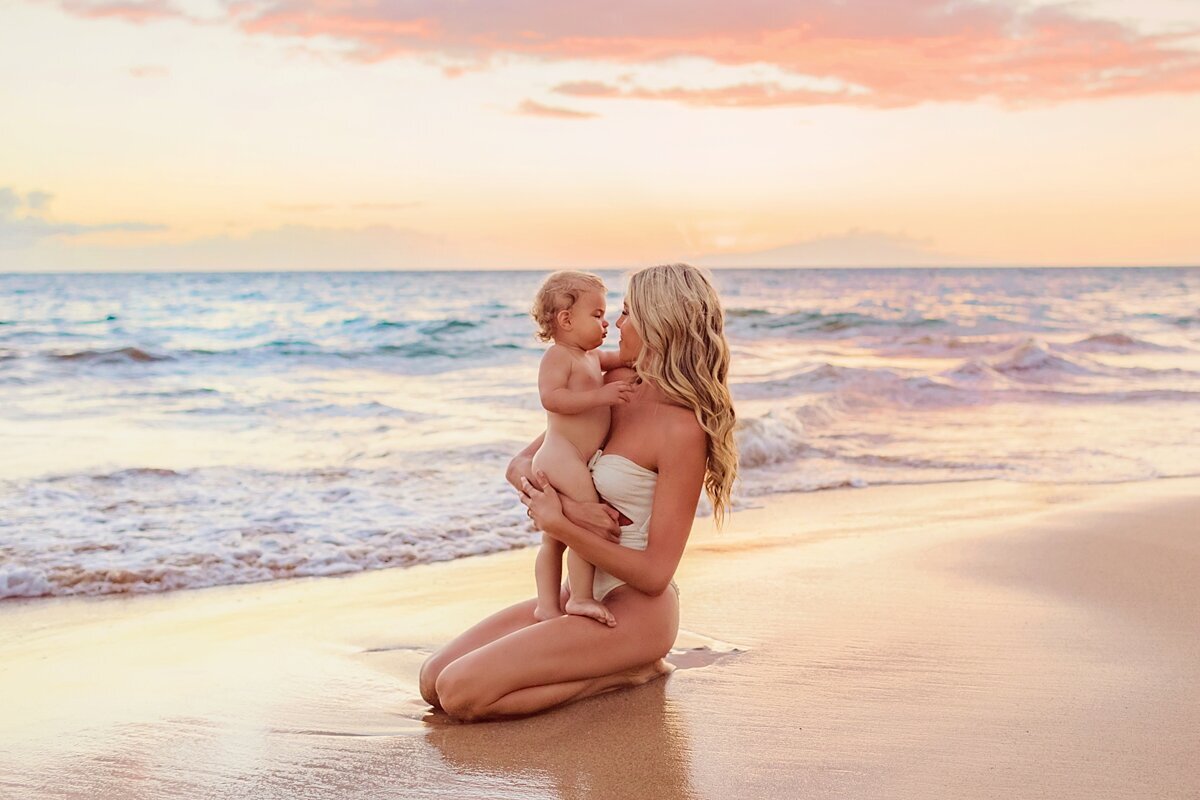 Mother with long blonde hair kneels on the sand and kisses her toddler just before sunset on the beach in Maui