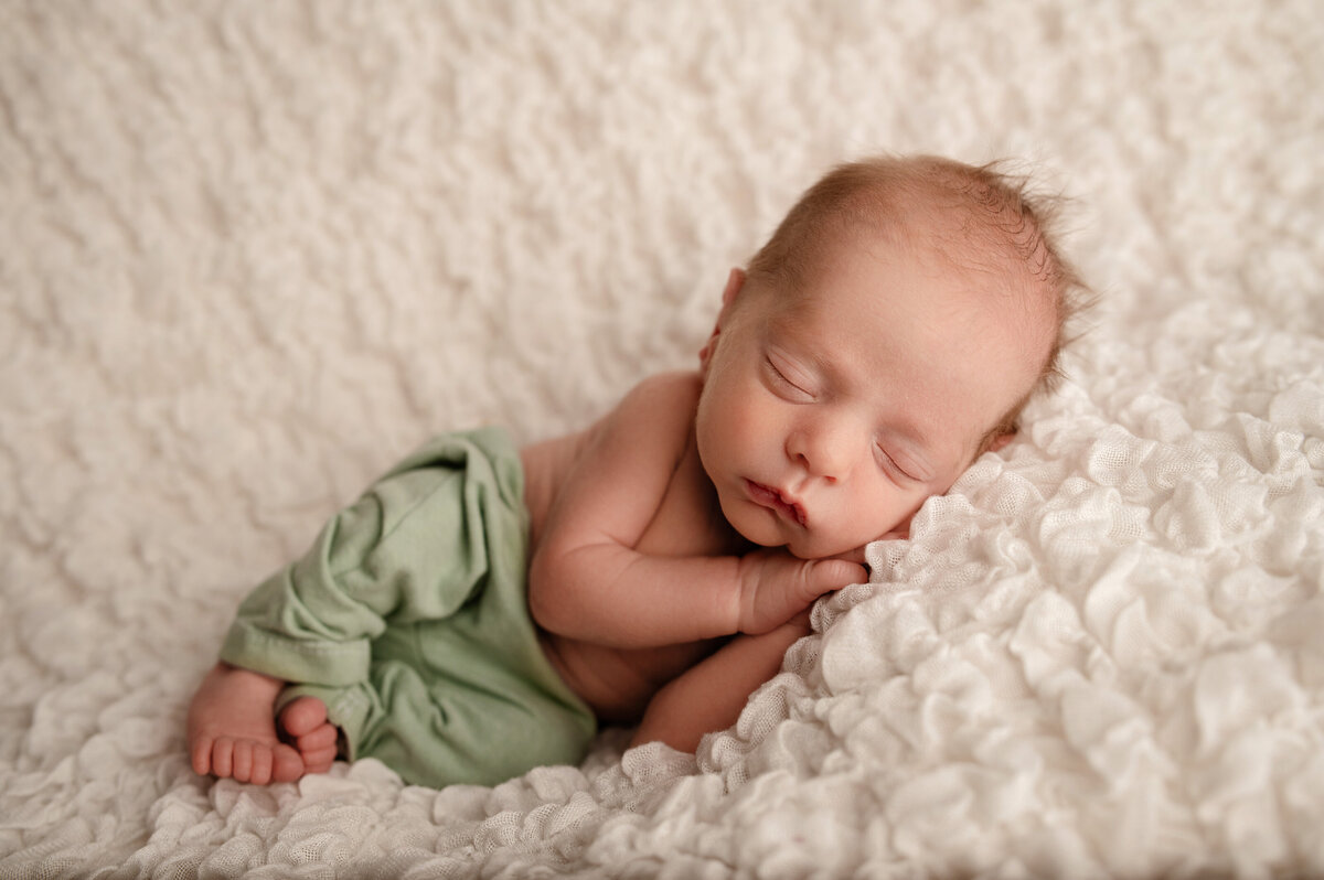 newborn boy laying on his side in sleepy pose wearing green pants and laying on a cream textured backdrop