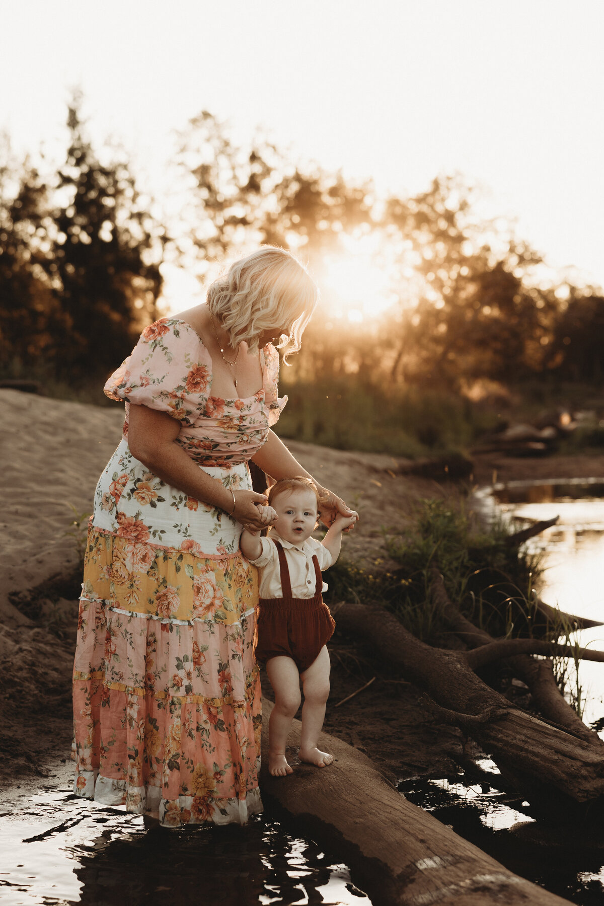 During a serene sunset, an adult helps a child stand on a log by the water's edge, capturing the essence of motherhood photography in Wollongong.