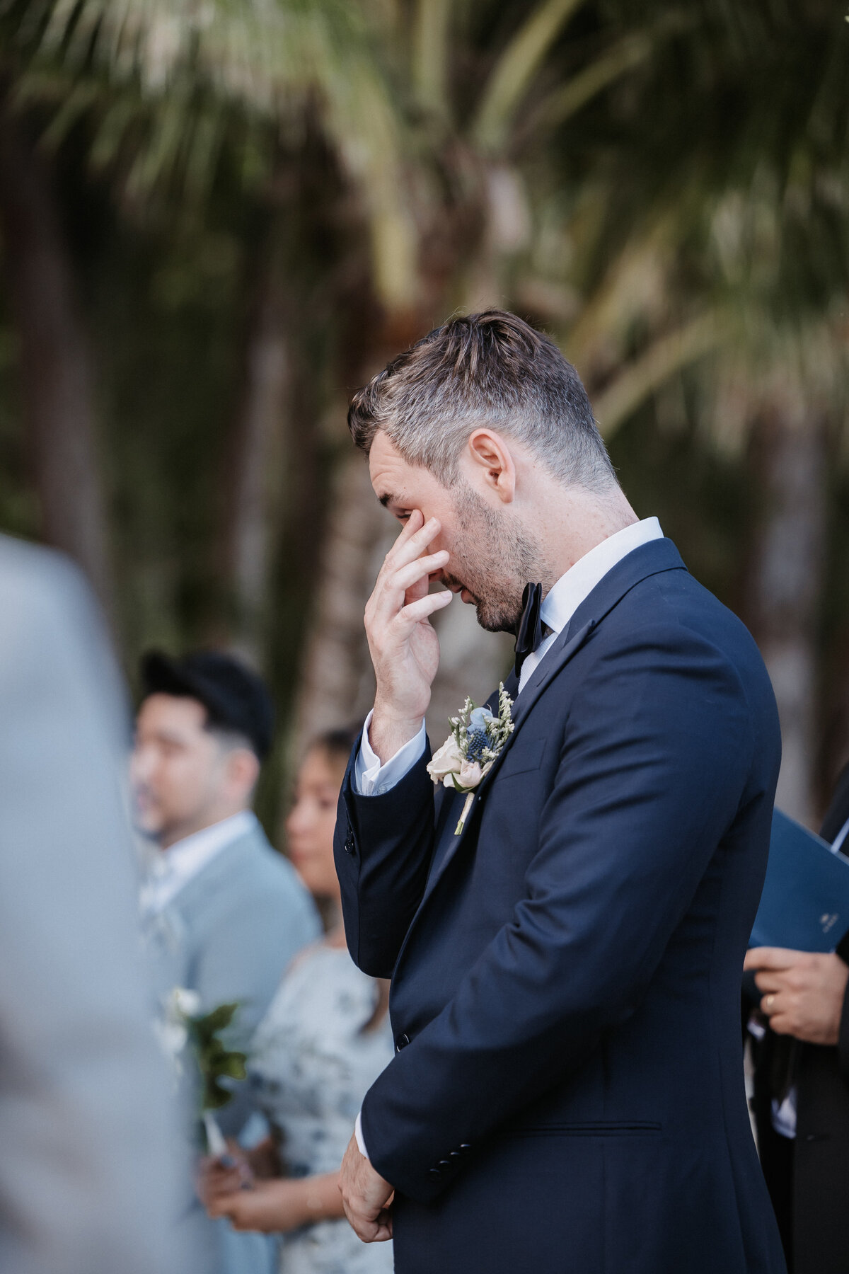 Emotional groom wiping tear from his eye wearing navy blue suit