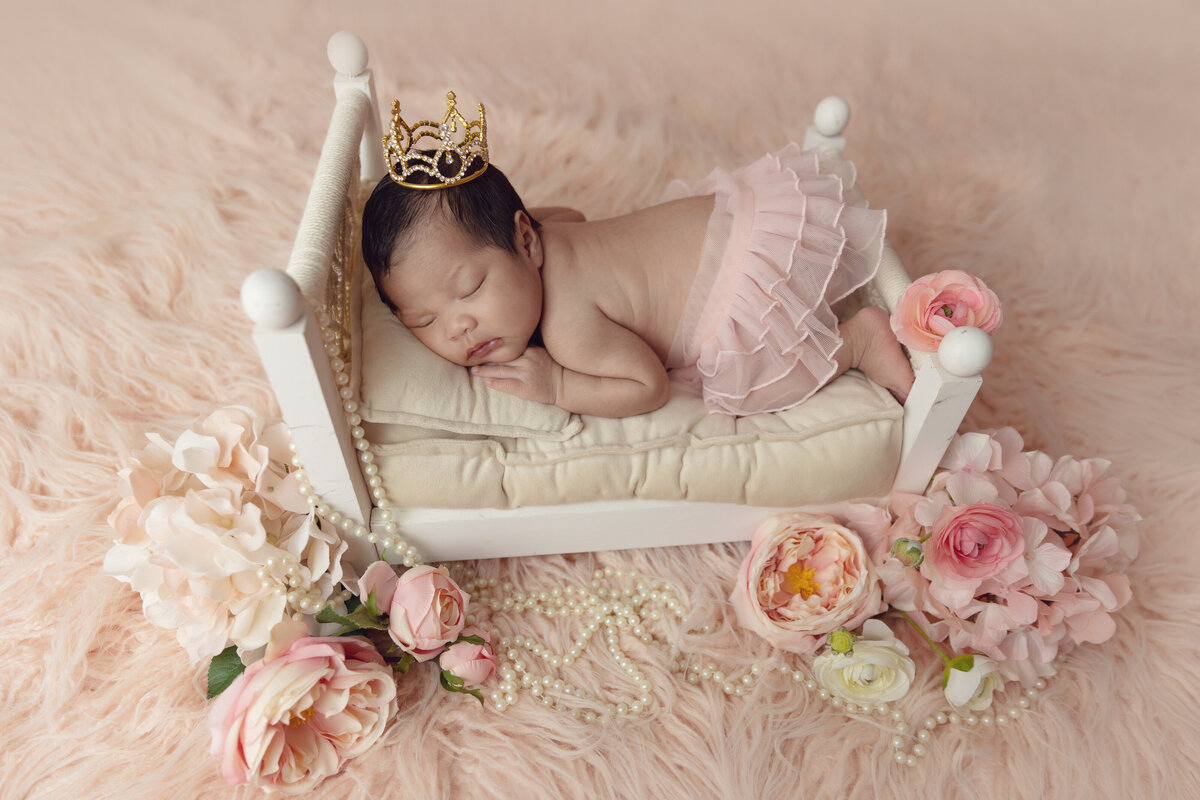 A newborn baby girl sleeps in a pink tutu on a wooden wed with flowers and pearls