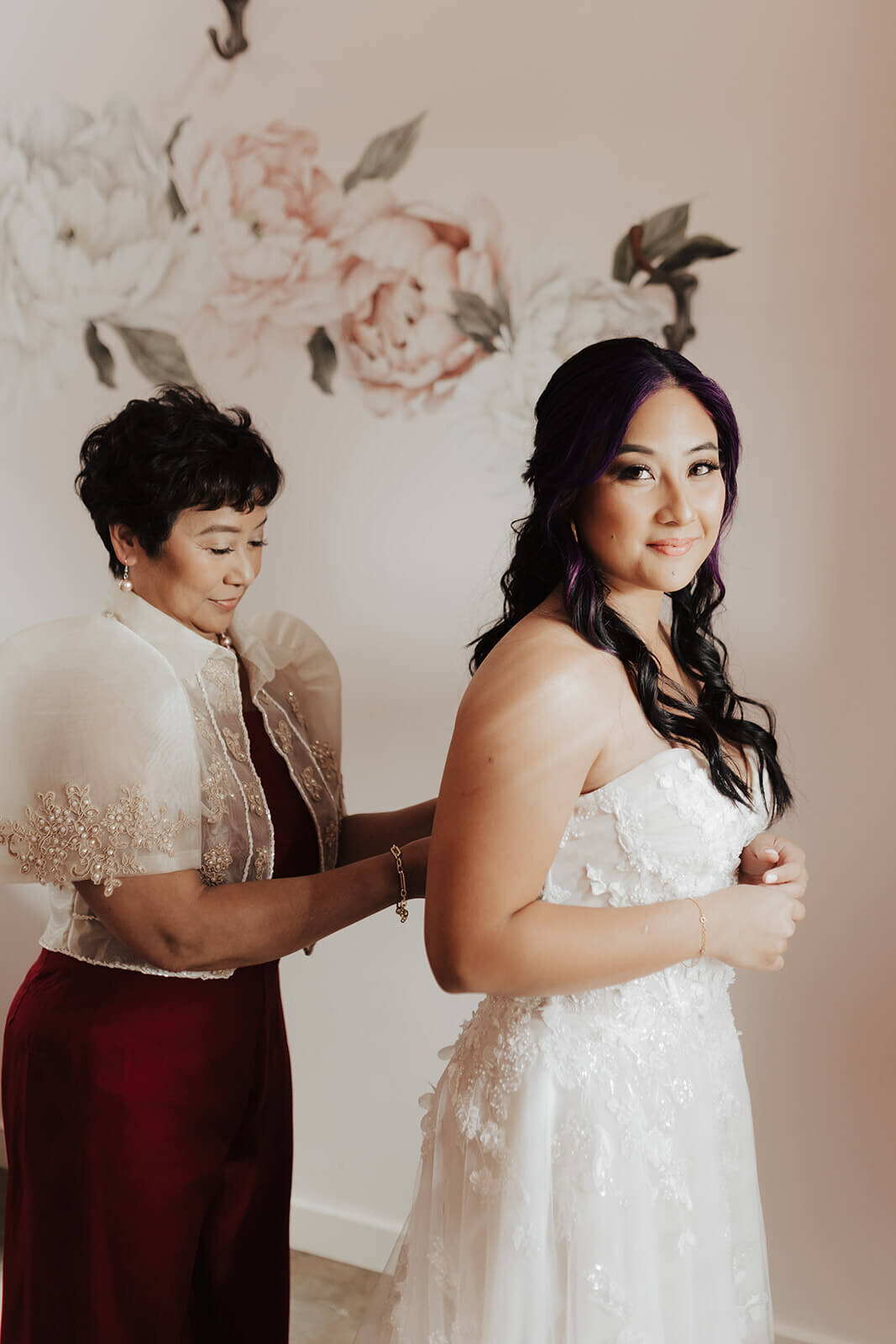 A beautiful bride with soft curls and radiant makeup smiles as her mother helps her into her wedding gown. Captured in a sweet getting-ready moment, this image highlights natural bridal makeup and soft romantic waves, perfect for a wedding day look. Ideal for brides in Austin or San Antonio seeking a mobile makeup artist or bridal hairdresser. Mistique Makeup specializes in wedding hair and makeup, offering on-location services for brides and wedding parties.