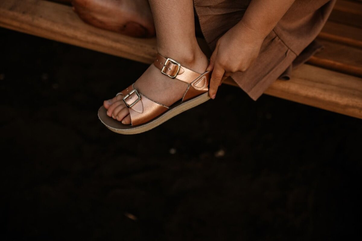 A person's hand adjusts the gold buckle of a Salt Water Sandals Surfer style sandal on their foot, while seated on a wooden surface. Close-up footwear shot