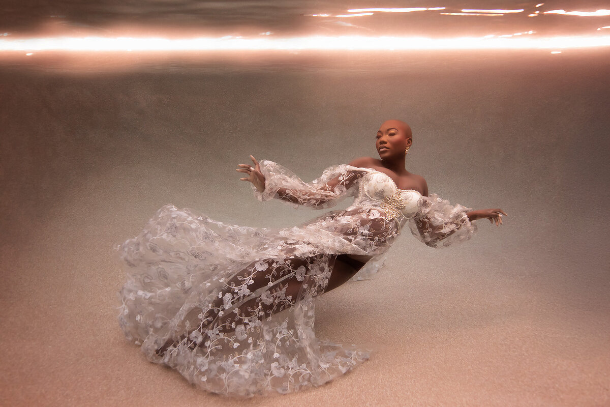 A serene underwater maternity photo showcasing a woman gracefully floating in a delicate, white lace gown with intricate floral patterns. The warm golden light reflects softly from the water's surface, creating a tranquil, dreamlike atmosphere. This Dallas underwater maternity photographer session captures the elegance and peaceful beauty of pregnancy in an artistic, underwater setting.