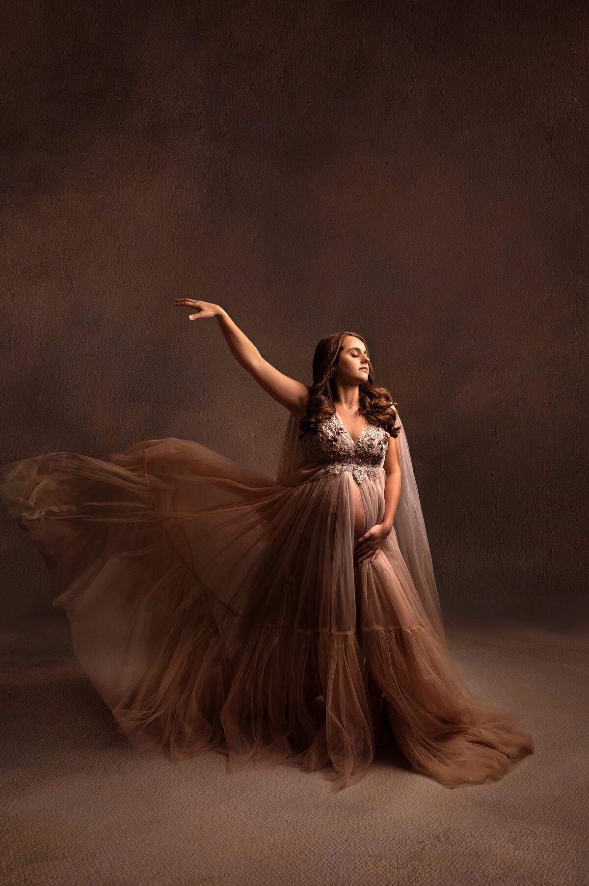 Pregnant woman wearing a flowing mocha tulle gown with embroidered bodice, captured mid-movement with a dramatic painterly backdrop in a Brookfield-area photography studio.