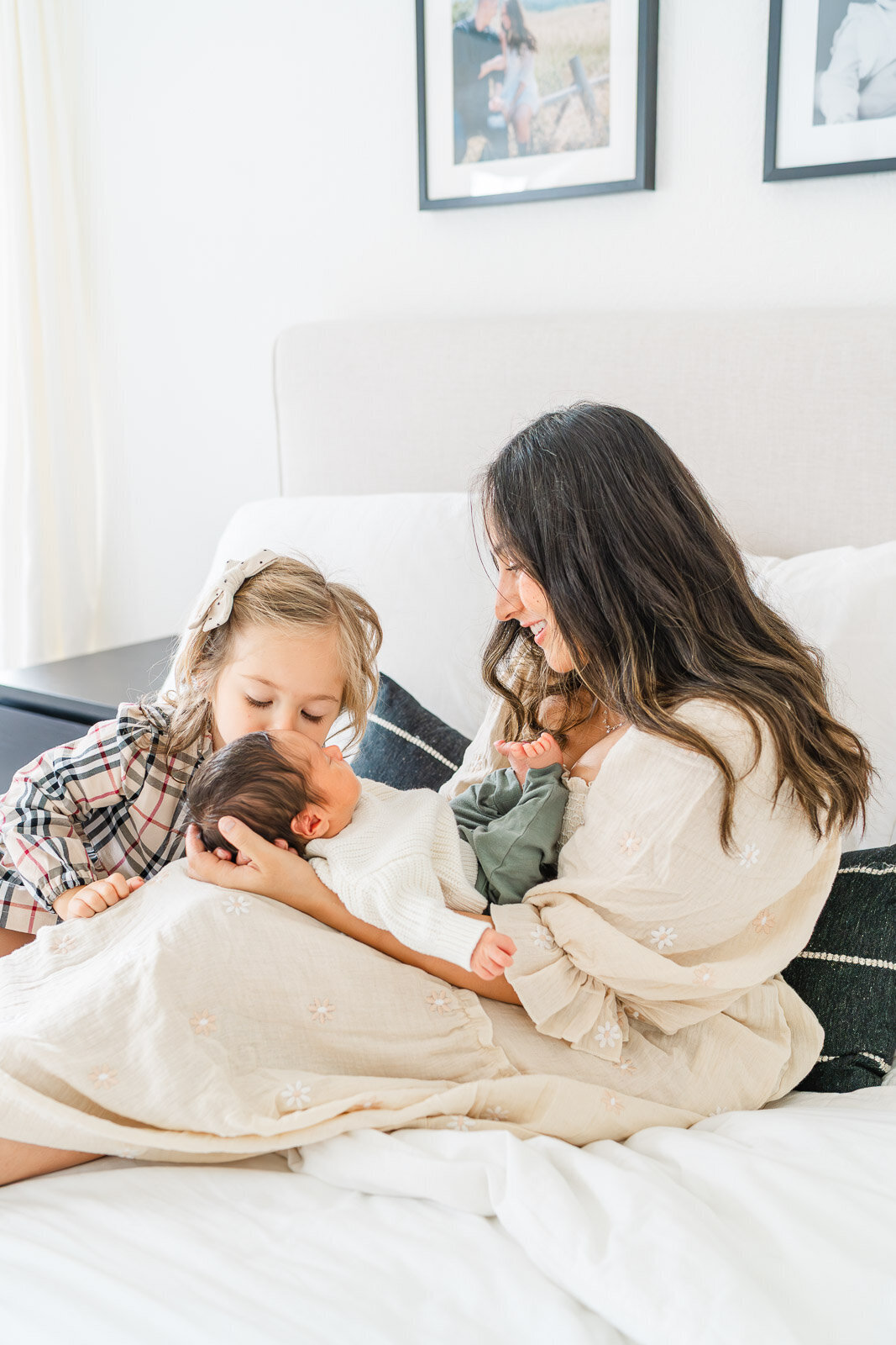 a mother holds her newborn while sitting on the bed. A toddler kisses the newborn on the forehead.