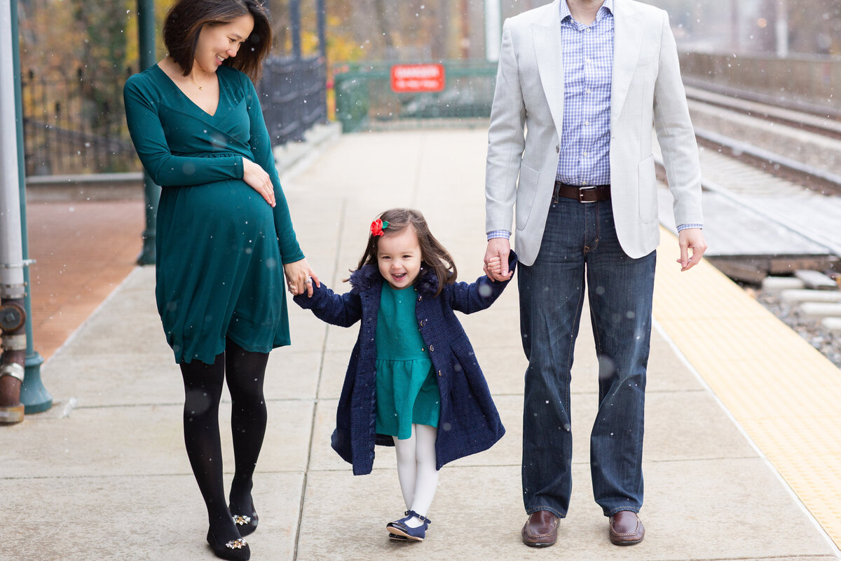 Winter maternity portrait of a couple with their young daughter in between, mom cradling her belly while snowflakes gently fall