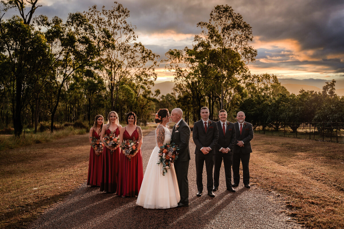 bride and groom sharing a kiss at sunset with bridal party behind them - Townsville Wedding Photography by Jamie Simmons
