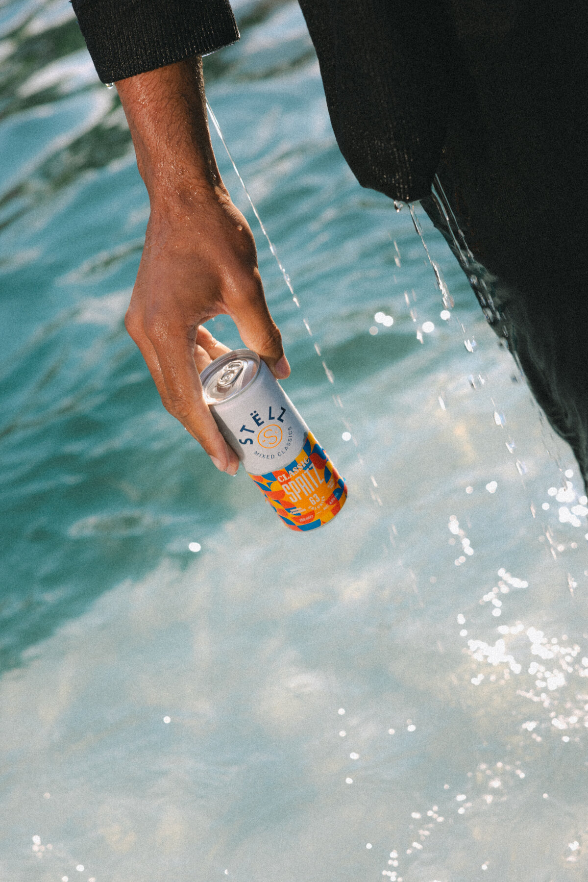 A close-up shot of a man's hand holding a STËLZ Classic Spritz can as water drips from his arm into the pool. The droplets and lighting create a dynamic and refreshing scene. Photography by Bo Eppink.