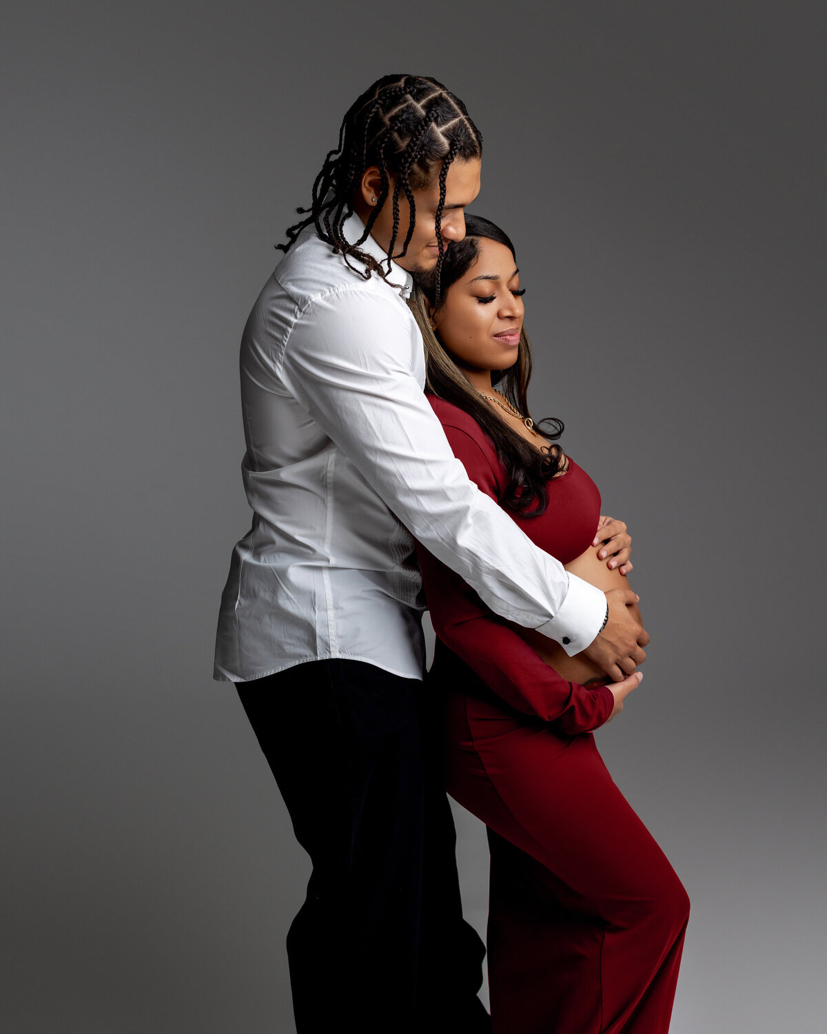 The mother-to-be in a red dress stands alongside her husband, both in a side view pose, with her lovingly holding her belly as they share a tender moment of anticipation. Captured by Inland Empire maternity photographer Kaitlyn Dawn Photography.