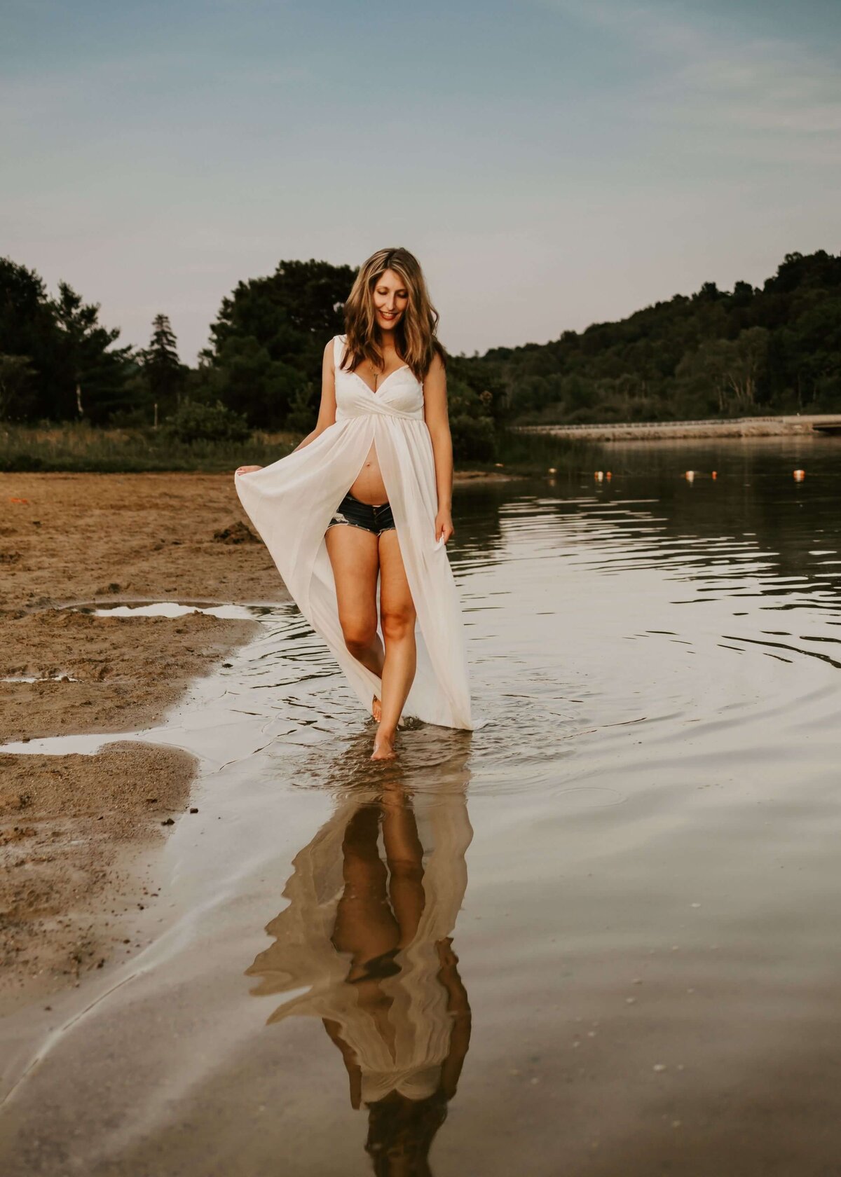 A woman in a white dress gracefully poses in the water for her Pittsburgh maternity photographer.