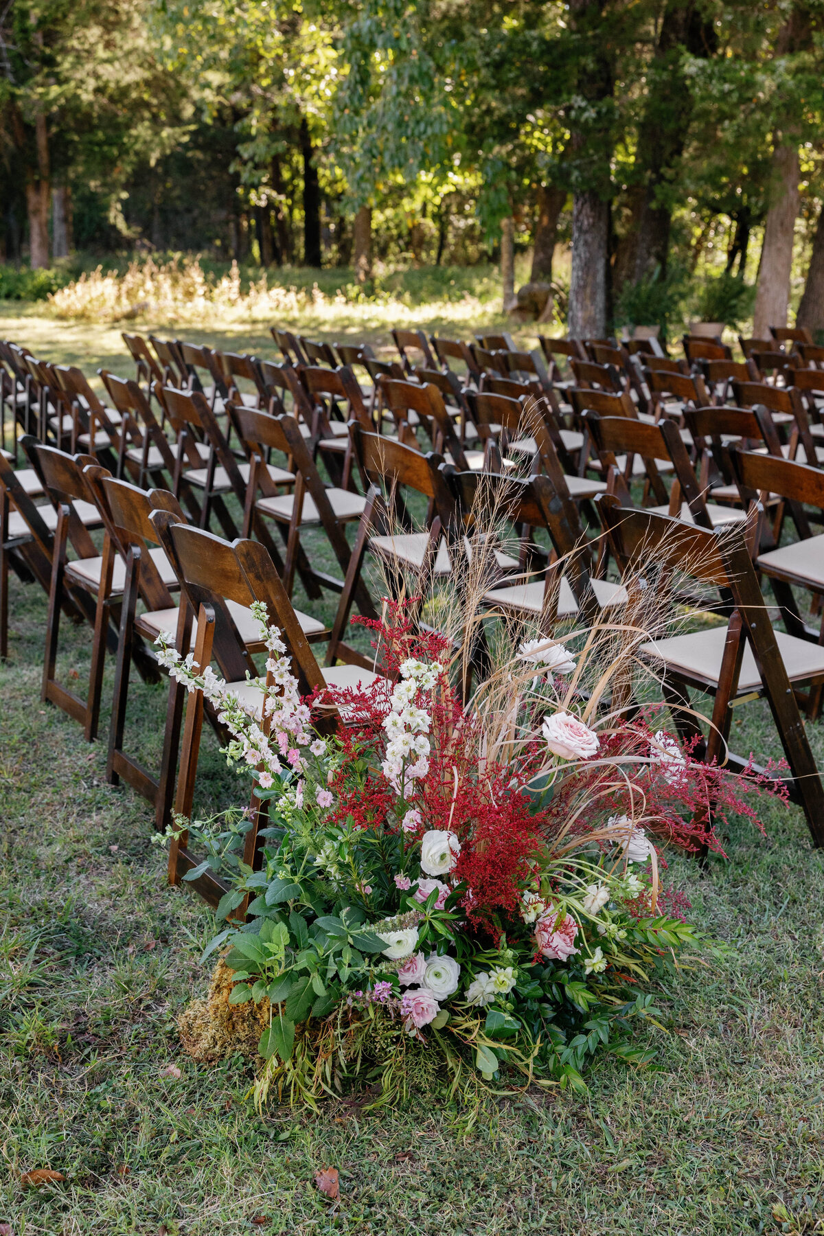 Lush ground floral arrangement of pink roses, red astilbe, grasses, and greenery lining the ceremony aisle at an outdoor Arkansas wedding, designed in an organic garden-inspired style.