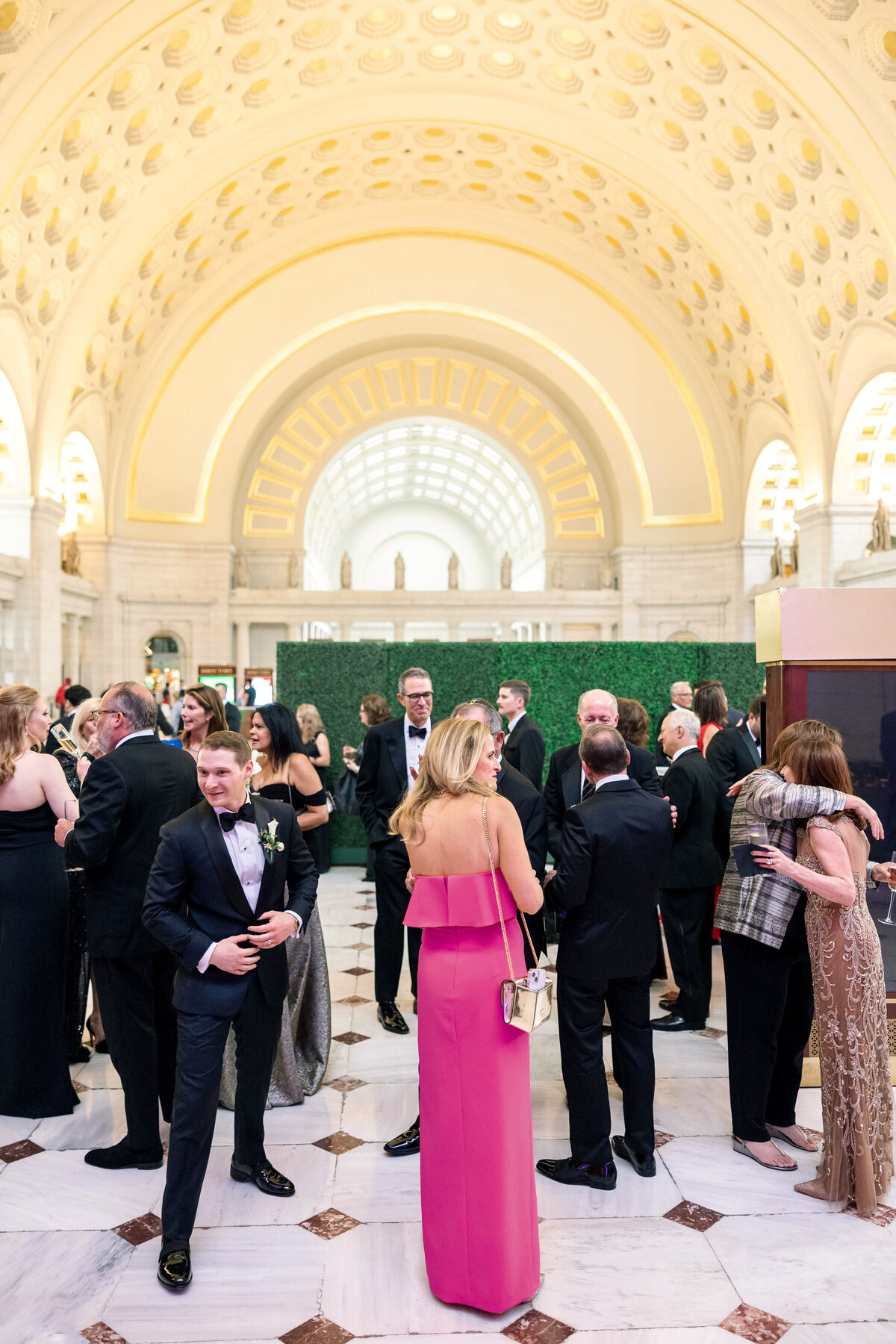 A formal event in a grand hall with an arched, ornate ceiling. Guests in evening attire are socializing, some standing and chatting, while others embrace. The space is warmly lit, featuring a mix of elegant dresses and suits.