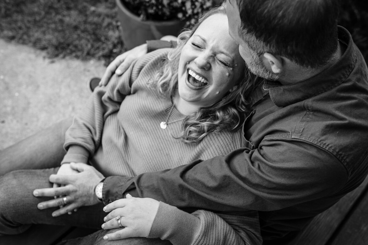A playful, close-up shot of a mother laughing heartily as her partner embraces her from behind, taken outdoors in black and white.