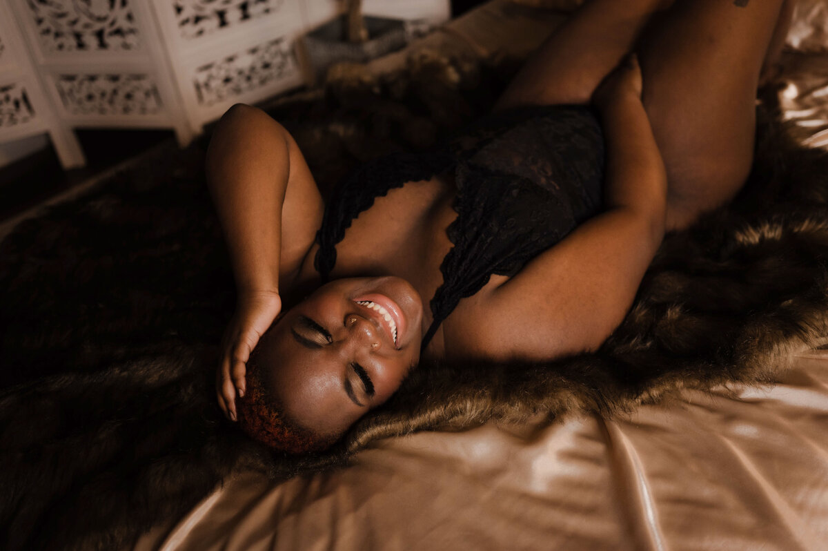 A woman in black lingerie lays on a bed laughing for a Memphis Boudoir Photographer