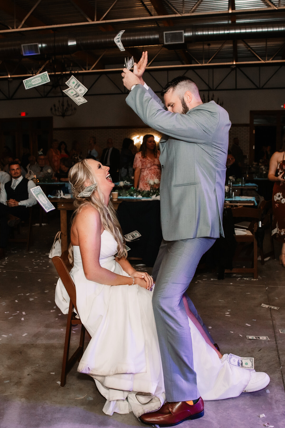 A bride sits in a wooden chair as her husband showers money over her and dances straddling her