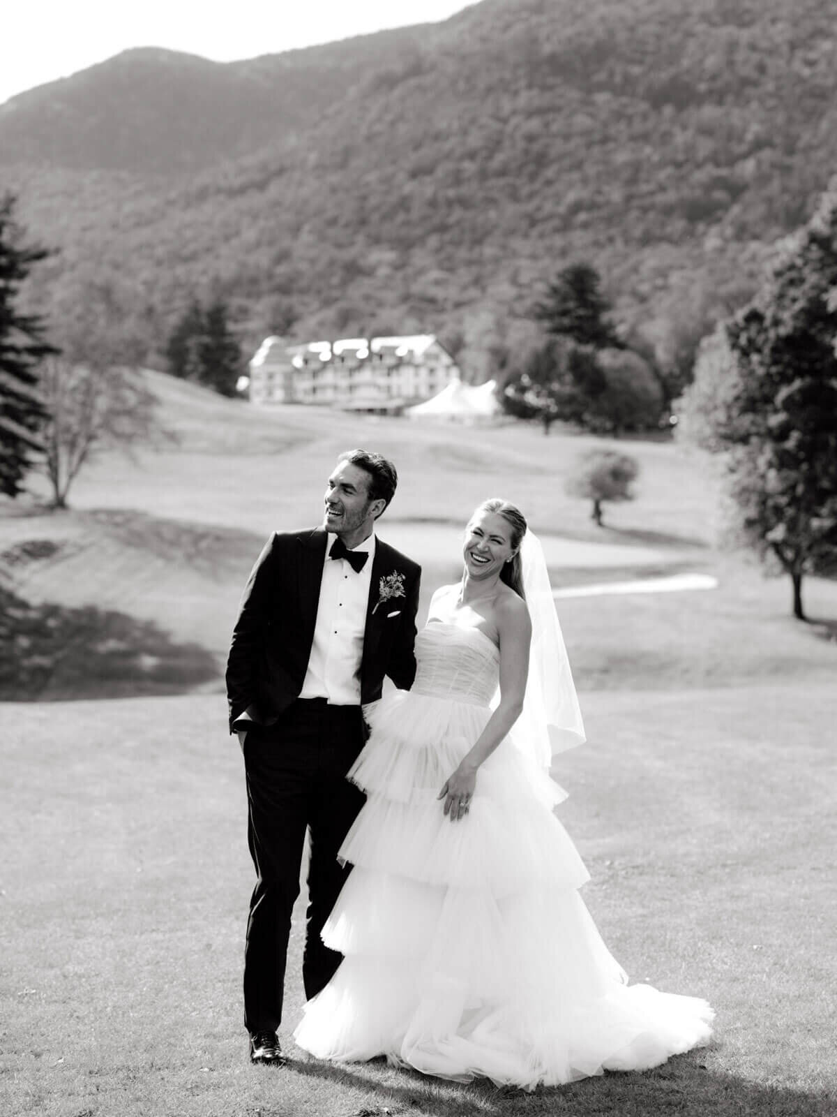 The bride is smiling at the camera, with the groom beside her, at The Ausable Club's golf course, NY. Image by Jenny Fu Studio.
