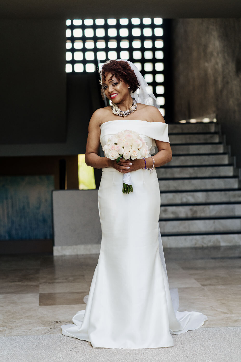 Bride in an off-the-shoulder gown holding a bouquet, standing by modern architecture