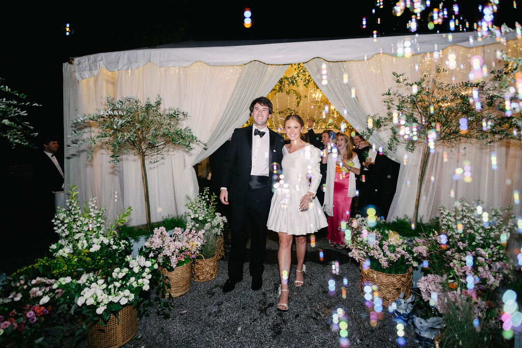 Bride and groom walk out of their tented reception at The Bascom in Highlands, NC during a romantic bubble exit surrounded by lush floral décor.