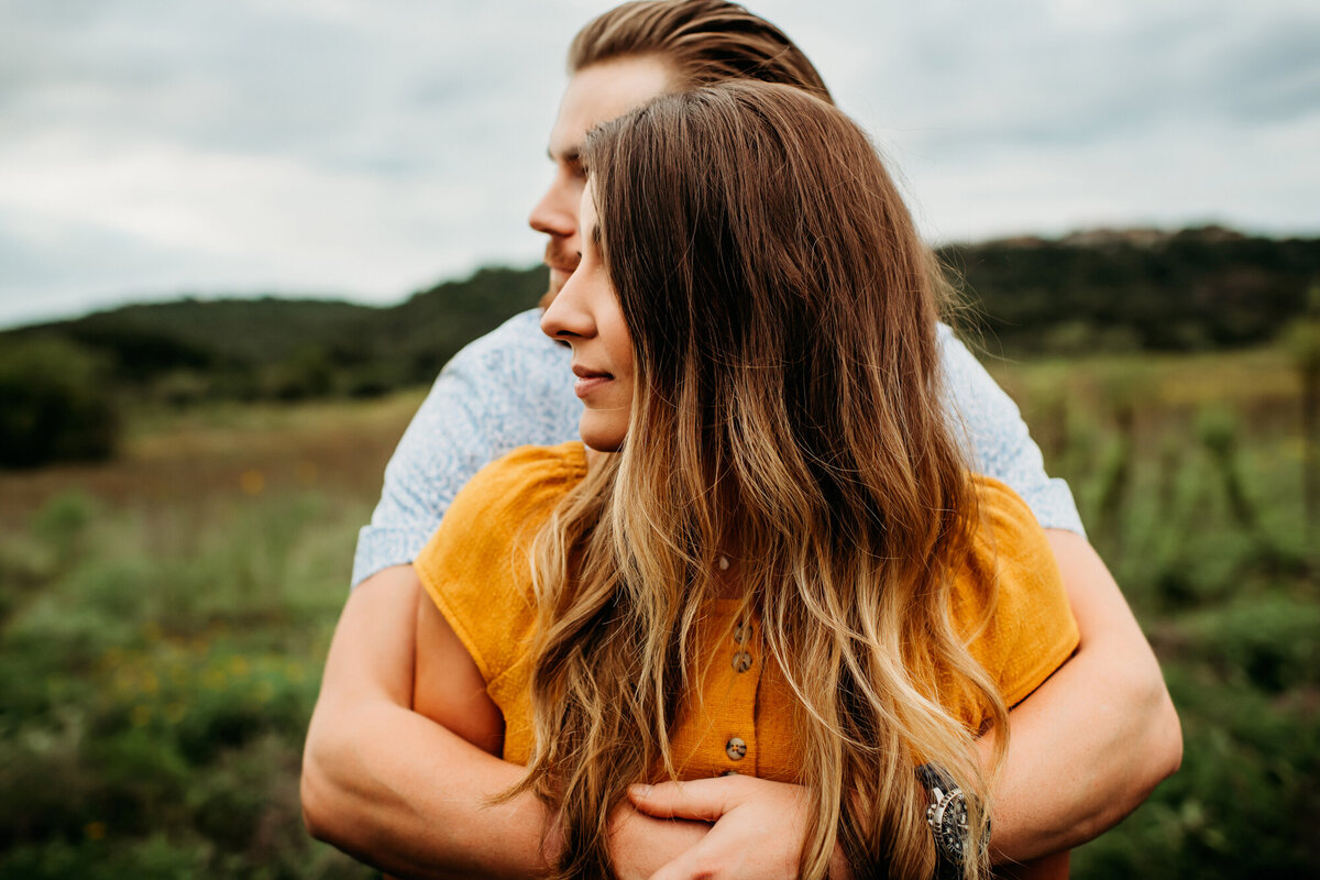 Couples Photography, Man hugs woman from behind, they stand in grassy field