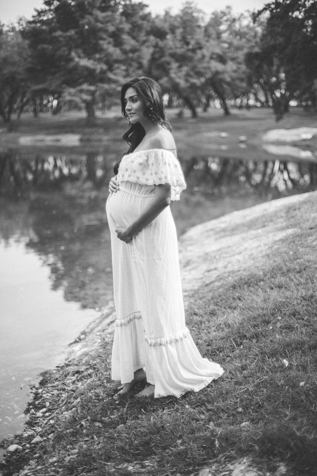 Maternity photography session of pregnant woman in white see through maternity dress standing next to lake.