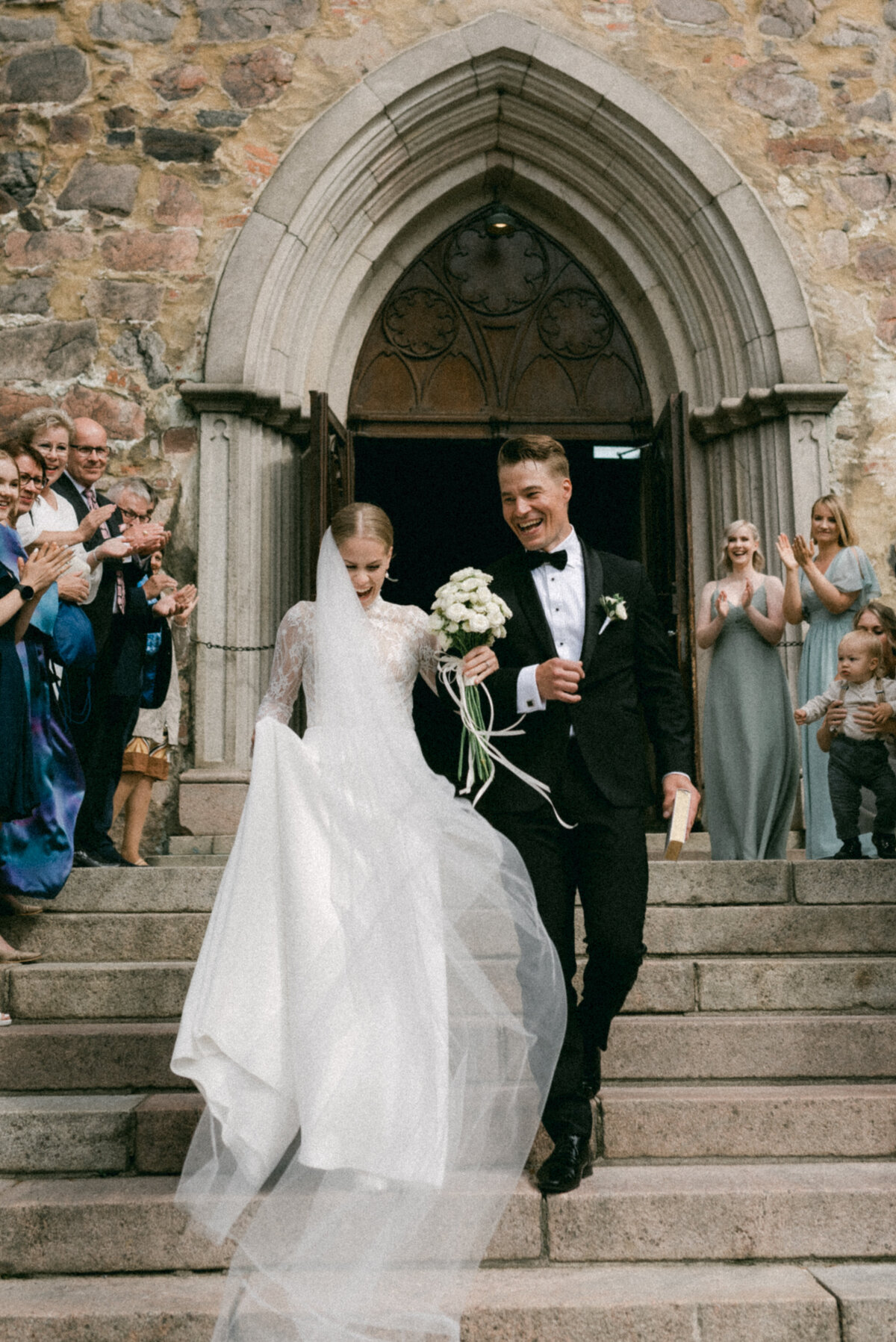Bride and groom walking out of the church after wedding ceremony. The weil is flying in the wind and the couple is laughing. Photograph captured by wedding photographer Hannika Gabrielsson.