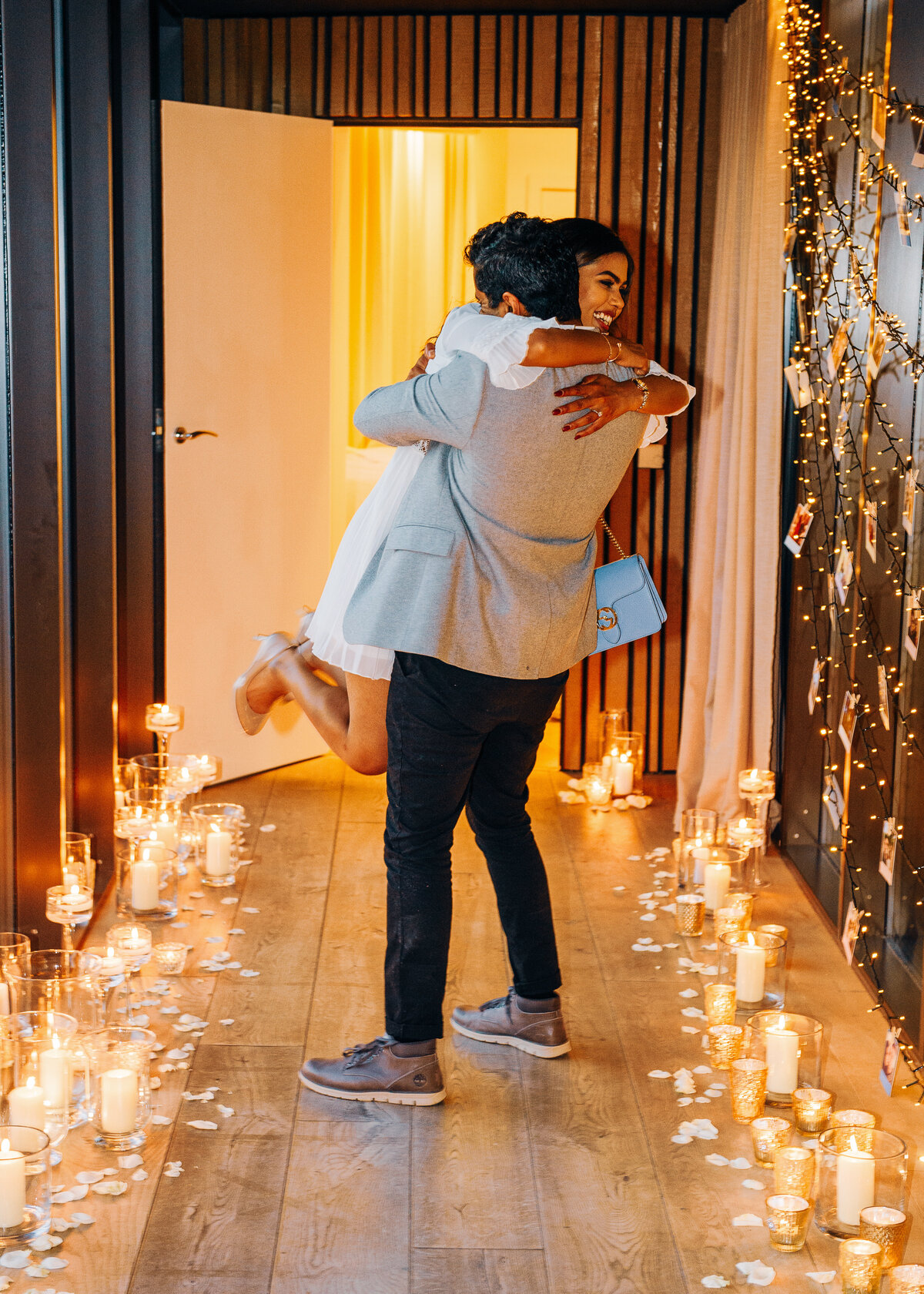 A couple embrace after getting engaged in a candlelit hallway with polaroids strung on fairy lights.