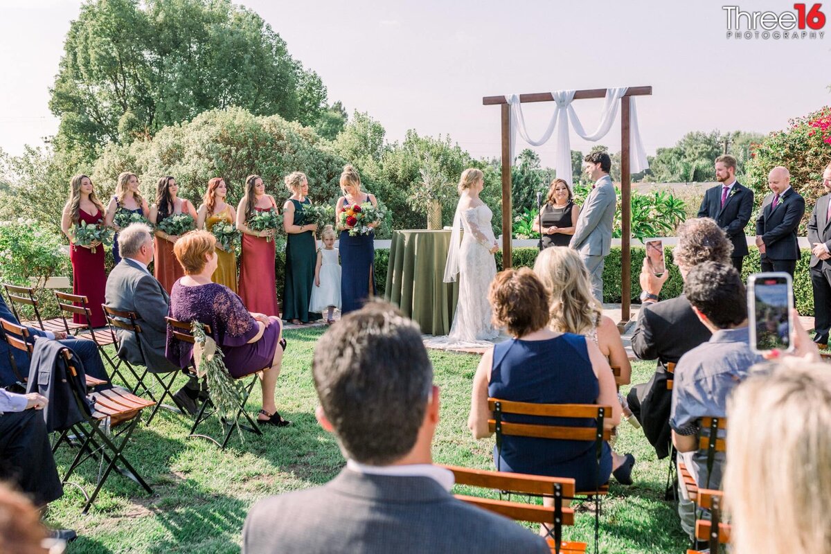 Bride and Groom stand at the altar facing each other during the ceremony in front of family, friends and wedding party