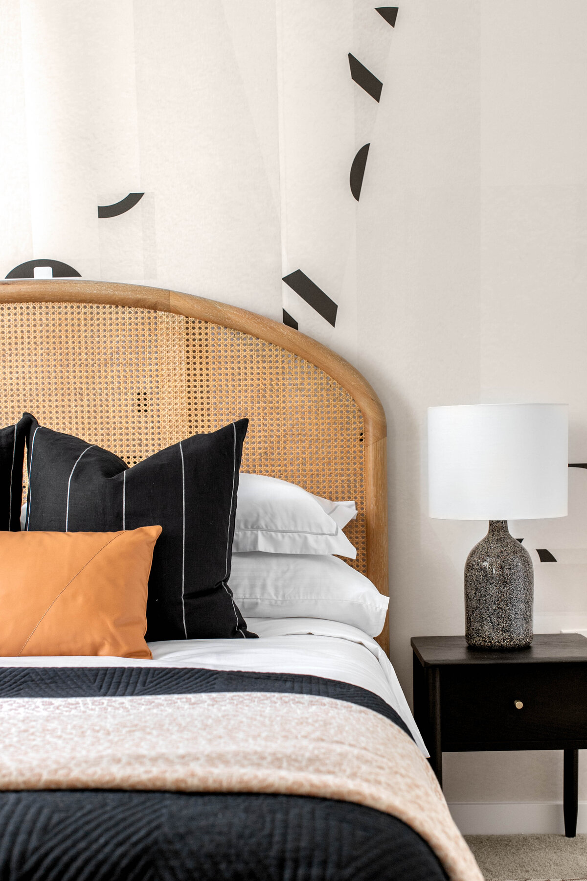 Bedroom featuring abstract geometric pattern wallpaper, cane bed headboard, striped black pillows, leather pillow and terrazzo bedside table lamp on top of black wooden single drawer nightstand